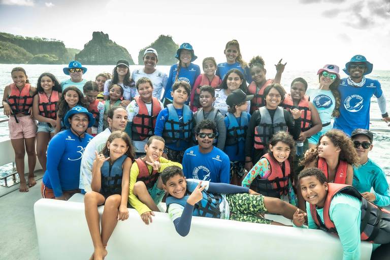 Um grupo de crianças e adultos posando em um barco， todos usando coletes salva-vidas. Eles estão sorrindo e fazendo poses divertidas. Ao fundo， é possível ver formações rochosas e o mar. A maioria das pessoas está vestindo camisetas de manga longa， algumas em tons de azul， e há um clima de alegria e descontração.