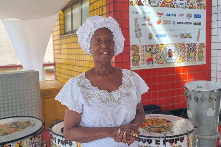 Uma mulher idosa está sorrindo em frente a um fundo colorido com azulejos. Ela usa um vestido branco com detalhes e um lenço na cabeça. Ao fundo， há tambores decorados e uma parede com cartazes e informações visíveis.