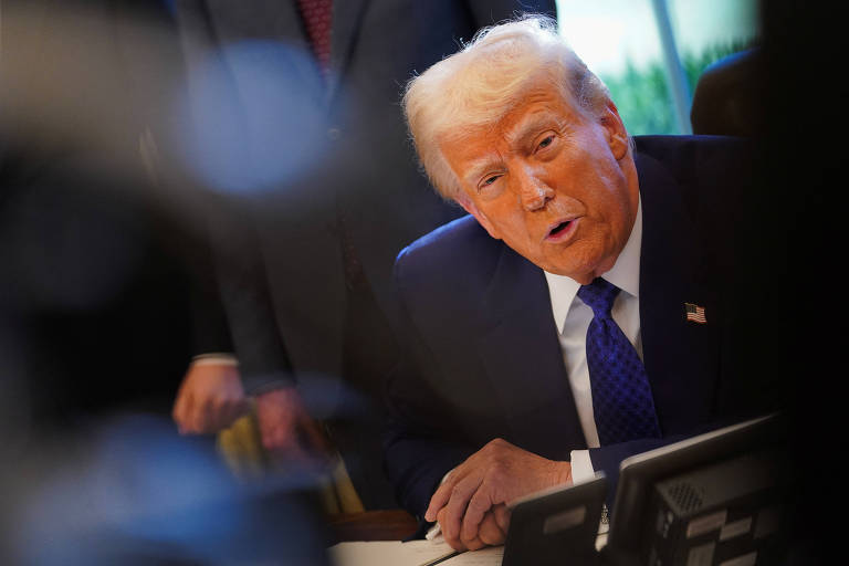 Trump， um homem branco， está sentado em uma mesa， olhando para a câmera. Ele usa um terno escuro e uma gravata azul. Ao fundo， há outras pessoas e um ambiente de escritório bem iluminado.