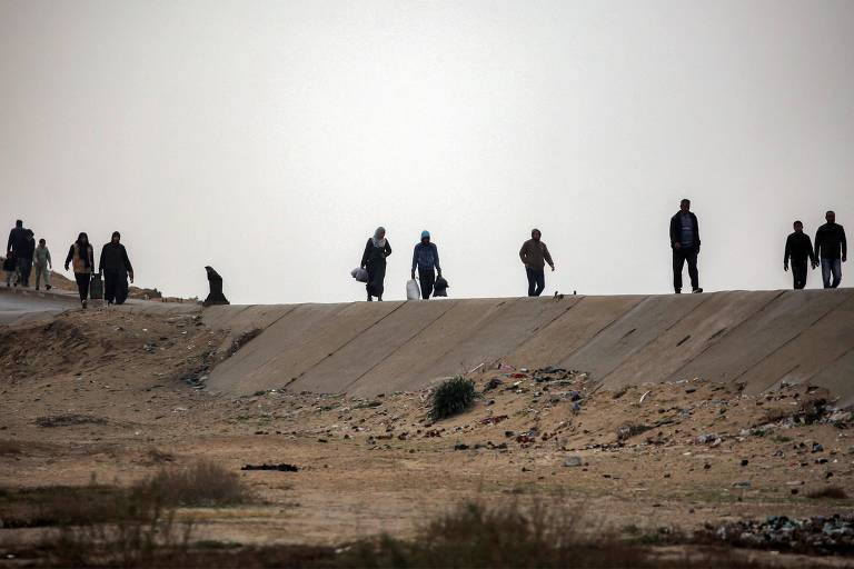 A imagem mostra várias pessoas caminhando em um terreno árido， com um caminho de concreto ao fundo. As figuras estão distantes e em silhueta， com algumas carregando objetos. O céu está nublado， criando uma atmosfera sombria.