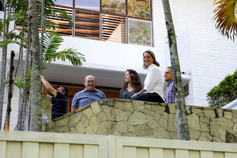 Cinco pessoas estão em uma varanda de uma casa moderna， com paredes brancas e grandes janelas. Elas estão em pé sobre uma parede de pedra， cercadas por plantas. A cena é iluminada pela luz natural do dia.