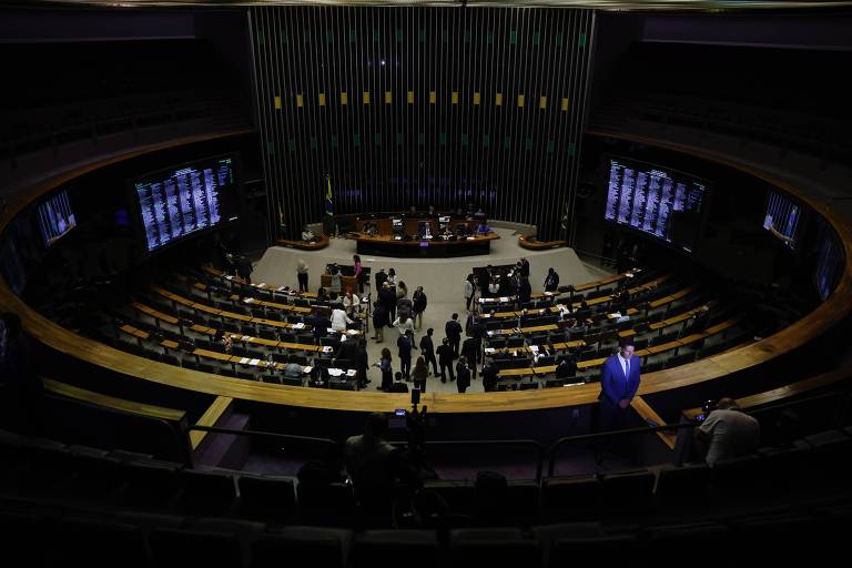 Imagem do plenário da Câmara dos Deputados do Brasil， com várias pessoas em pé e sentadas， algumas em grupos conversando. O ambiente é circular， com assentos dispostos em fileiras e uma mesa central onde estão alguns representantes. Telas de exibição estão visíveis nas laterais， e a iluminação é suave.