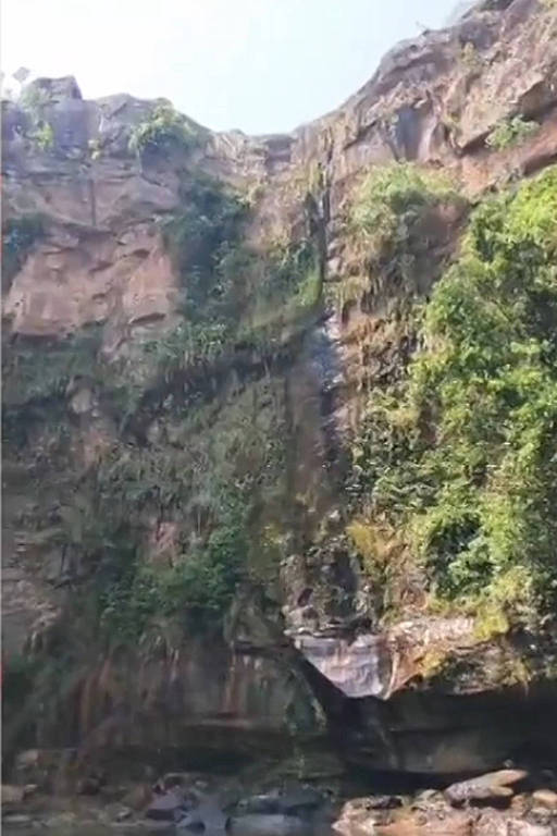 A imagem mostra uma cachoeira caindo de um penhasco alto， cercada por rochas e vegetação densa. A água desce em um fluxo contínuo， formando uma pequena piscina na parte inferior， onde algumas pedras estão visíveis. O céu está claro， e a luz do sol ilumina a cena.