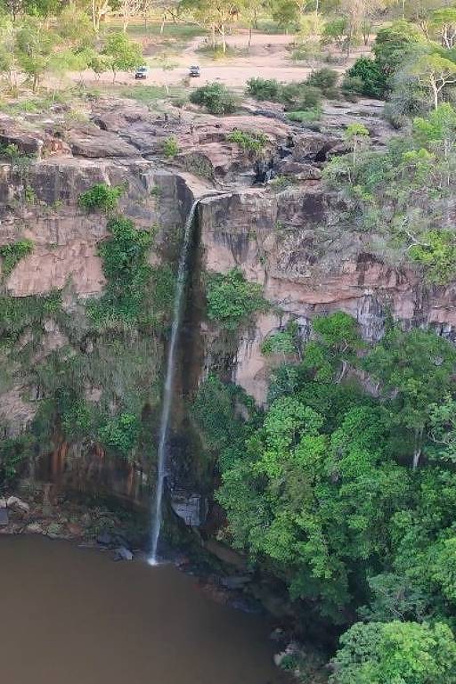 A imagem mostra uma cachoeira que deságua em um lago cercado por vegetação densa. As rochas ao redor são de cor marrom e há árvores verdes ao redor da área. O céu é claro e a paisagem é natural, com uma sensação de tranquilidade.