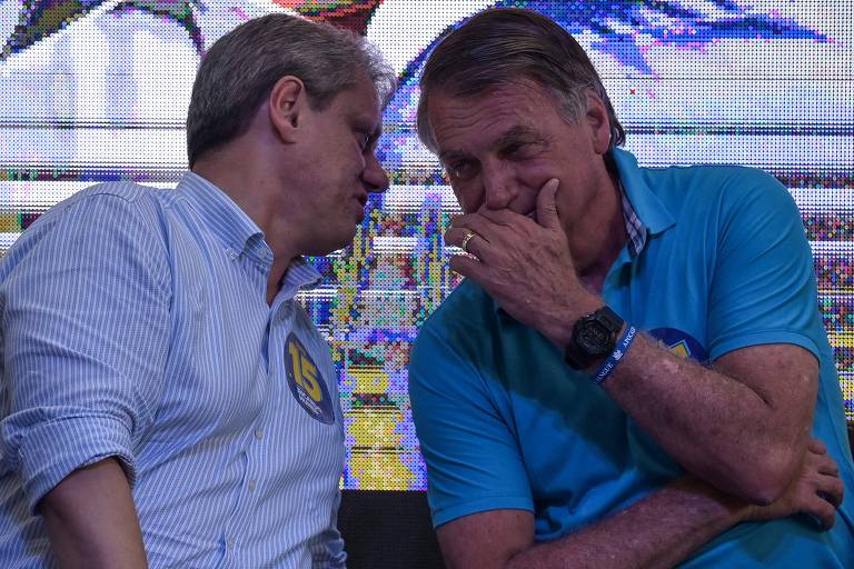 Dois homens estão sentados próximos um do outro， conversando em um ambiente informal. Um deles usa uma camisa branca e o outro uma camisa azul. Ambos parecem estar em um momento de descontração， com expressões faciais que indicam interesse na conversa.
