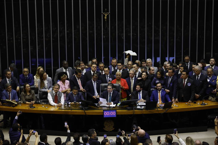 A imagem mostra uma sessão na Câmara dos Deputados do Brasil， com vários deputados reunidos em torno da mesa. No centro， um deputado está falando， enquanto outros observam. Há uma grande quantidade de pessoas na plateia， algumas segurando celulares. Ao fundo， há uma cruz e um painel com um cronômetro marcando 00:00.