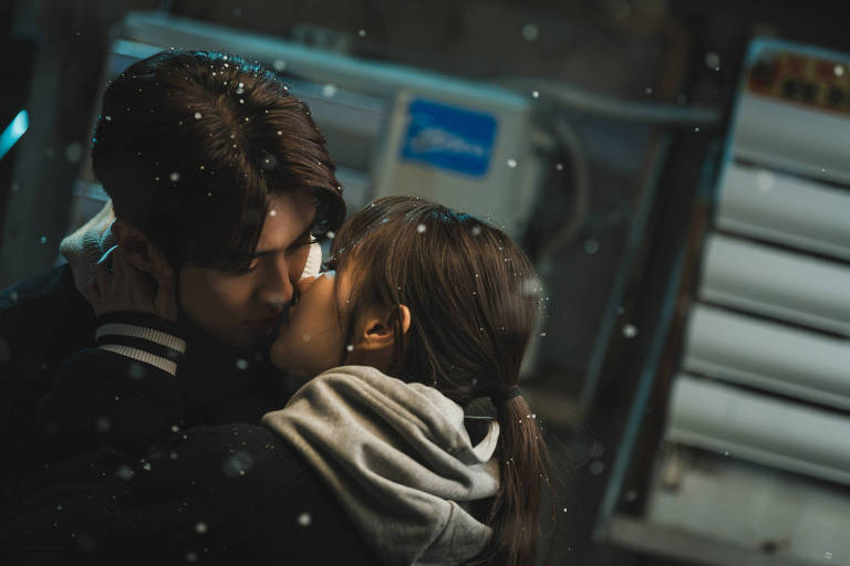 A imagem mostra um casal se beijando em um ambiente com neve caindo. Eles estão próximos um do outro， com expressões de carinho. O fundo é desfocado， mas há elementos que sugerem um ambiente urbano， como uma estrutura metálica e luzes. Ambos estão vestidos com roupas quentes， e a atmosfera é romântica.