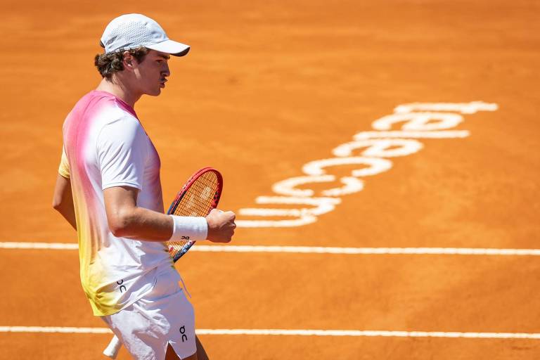 Um jogador de tênis está em uma quadra de saibro laranja， vestindo uma camiseta com gradiente de cores rosa e amarelo， e calças brancas. Ele segura uma raquete de tênis e parece estar comemorando um ponto. Ao fundo， há uma marcação no chão com a palavra 039;telecom039;.