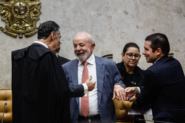 A imagem mostra um grupo de pessoas em um ambiente formal， possivelmente no Congresso. Um homem de terno claro e gravata rosa está sorrindo e interagindo com outros indivíduos. Um dos homens usa uma toga preta， enquanto uma mulher com óculos observa a cena. O fundo é decorado com um emblema， e as cadeiras são de couro marrom.