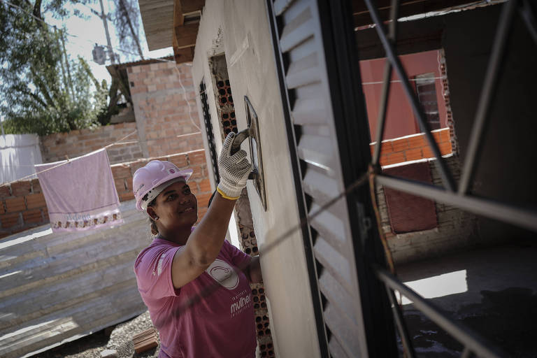 A imagem mostra uma mulher usando uma camiseta rosa e um capacete rosa， aplicando argamassa em uma parede de uma construção. Ela está concentrada no trabalho， com uma espátula na mão. Ao fundo， há uma área de construção com tijolos expostos e uma roupa pendurada para secar. A cena é iluminada pela luz do dia.