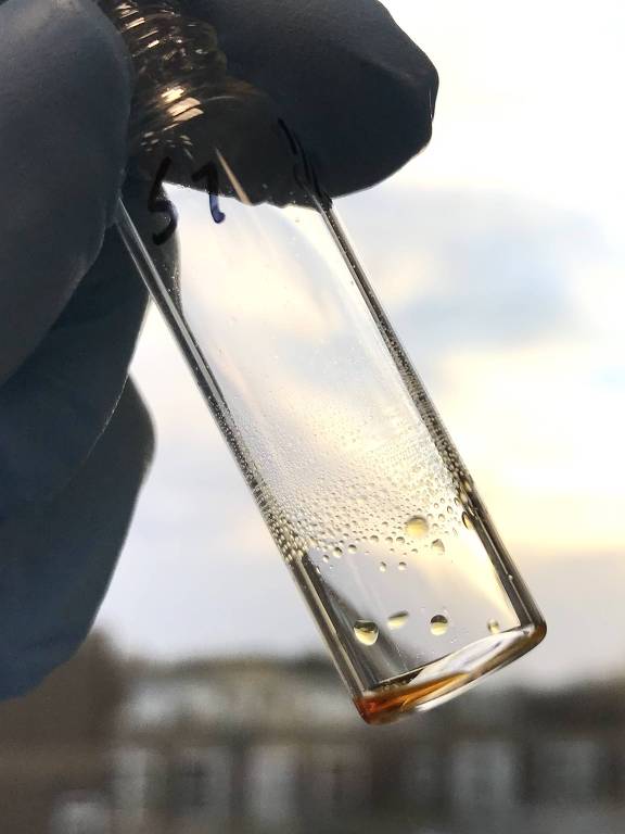 A imagem mostra um frasco de vidro transparente sendo segurado por uma mão com luva. O frasco contém um líquido claro com pequenas bolhas de ar. Ao fundo， há um ambiente desfocado， possivelmente um laboratório ou uma área externa com luz natural.