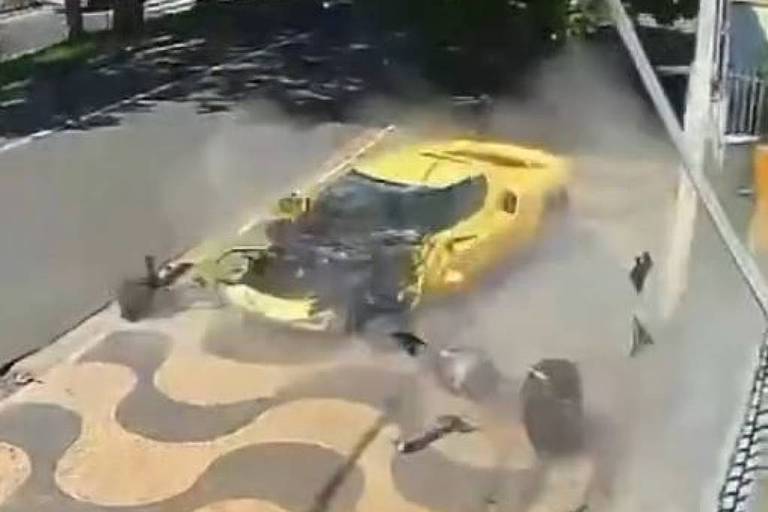 A imagem mostra um carro amarelo colidindo com a calçada em uma via pública. O carro está em movimento， levantando poeira e detritos ao redor.O cenário é ensolarado， com árvores ao fundo e um padrão de piso na calçada.