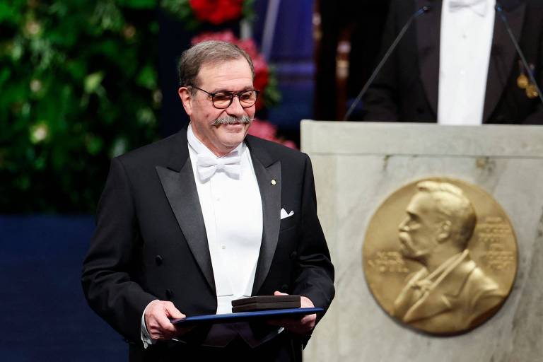 Um homem de terno preto e gravata borboleta branca segura um prêmio em uma cerimônia de premiação. Ele tem óculos e um bigode. Ao fundo， há uma mesa com um prêmio Nobel em destaque e um fundo decorado com plantas.