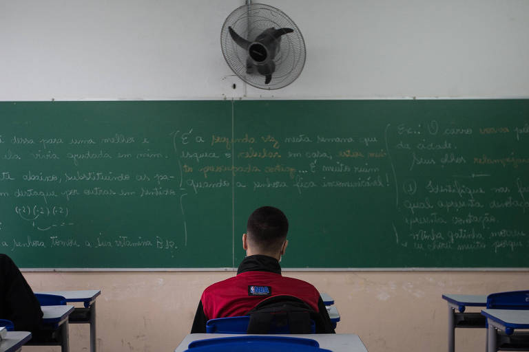 A imagem mostra uma sala de aula com mesas e cadeiras azuis. No fundo， há um quadro negro com texto escrito. Uma professora， usando máscara， está de pé em frente ao quadro， enquanto três alunos estão sentados， voltados para ela. A sala é bem iluminada e possui uma ventilador de teto.