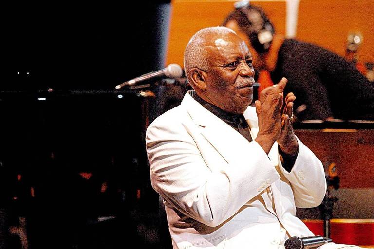 Um homem de pele escura， usando um terno branco e uma camisa preta， está aplaudindo enquanto sorri. Ele está em um palco， com um microfone à sua frente. Ao fundo， é possível ver instrumentos musicais e outros músicos. A atmosfera parece ser de uma apresentação ao vivo.