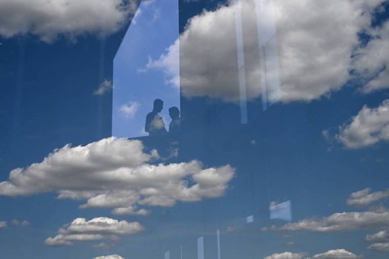 A imagem mostra um céu azul com nuvens brancas. No primeiro plano， há reflexos de duas pessoas em uma superfície de vidro， que se sobrepõem ao céu. As nuvens estão dispersas e o ambiente parece ser iluminado pelo sol.