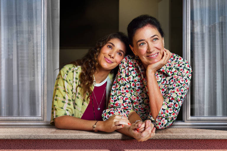 Duas mulheres estão sorrindo e posando juntas em uma janela. Elas estão usando roupas coloridas e estampadas. A parede ao redor da janela é de cor rosa， e há cortinas brancas visíveis. O céu pode ser visto ao fundo， através da janela.