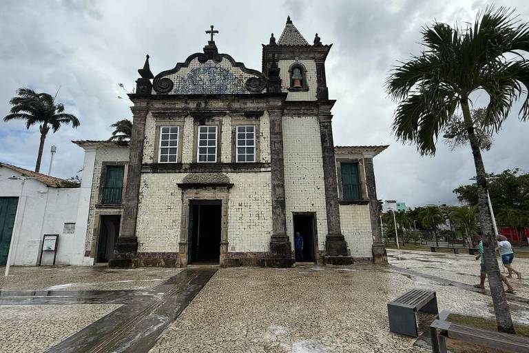 A imagem mostra uma igreja de estilo colonial， com uma fachada de azulejos brancos e detalhes em pedra. A estrutura possui uma torre com um pequeno cruz e janelas grandes. À frente， há um calçamento de pedras e algumas palmeiras ao redor. O céu está nublado， indicando um dia chuvoso.