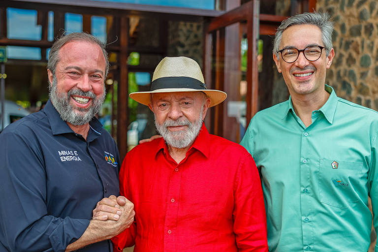 Três homens estão posando juntos em frente a um edifício com paredes de pedra. O homem à esquerda tem cabelo e barba grisalhos， usa uma camisa preta com o logo de uma empresa e está segurando a mão do homem do meio. O homem do meio， que usa um chapéu claro e uma camisa vermelha， sorri. O homem à direita tem cabelo grisalho e usa óculos， vestindo uma camisa verde. Ao fundo， há janelas de madeira e uma vista de natureza.