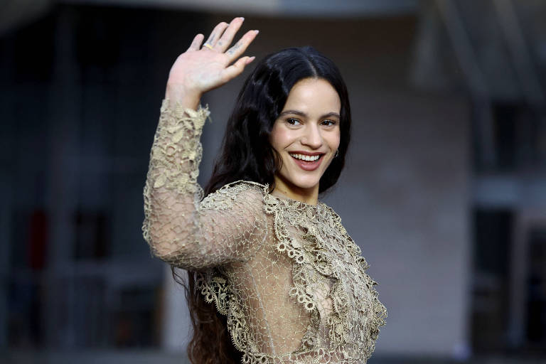Uma mulher com cabelo longo e escuro, vestindo um vestido transparente com detalhes em renda, está acenando com a mão direita e sorrindo. O fundo é desfocado, sugerindo um ambiente ao ar livre ou um evento.