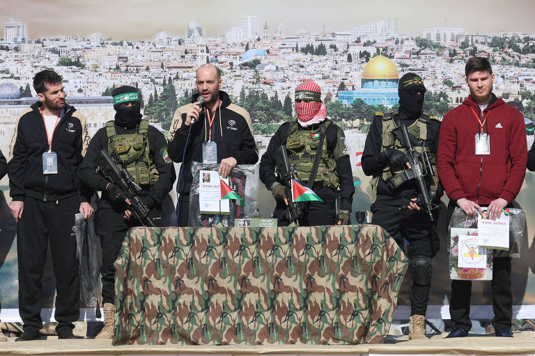 A imagem mostra um grupo de seis homens em um evento. Quatro deles estão vestidos com uniformes militares e armados， enquanto um deles está falando ao microfone. O homem ao centro usa uma camisa preta e está cercado por outros que usam máscaras e lenços. Ao fundo， há uma imagem de uma cidade com uma cúpula dourada visível. Uma mesa camuflada está à frente， com bandeiras da Palestina e documentos.