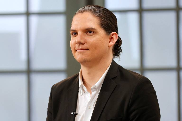 Um homem com cabelo preso em um coque， usando um paletó preto e uma camisa branca， está sentado em um estúdio. Ele tem uma expressão neutra e está olhando para frente. Ao fundo， há janelas grandes com luz natural entrando.