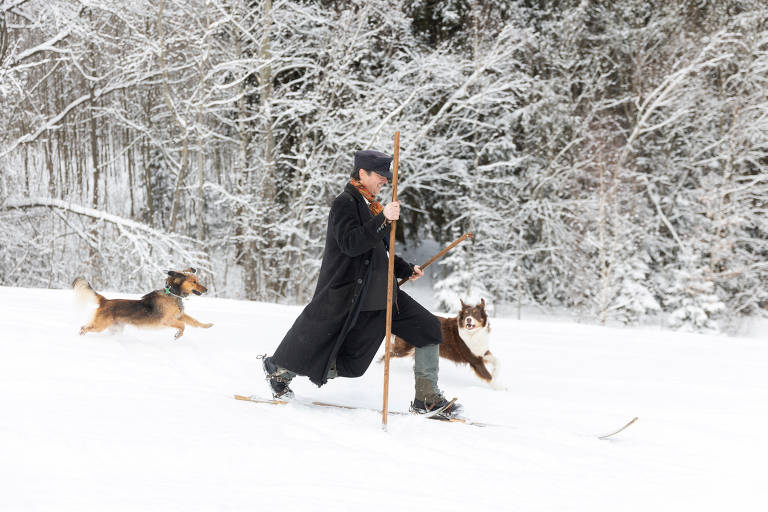 Um homem vestido com um casaco longo e chapéu está esquiando na neve, usando um bastão. Ele é acompanhado por dois cães que correm ao seu lado. O cenário é uma paisagem de inverno com árvores cobertas de neve ao fundo.