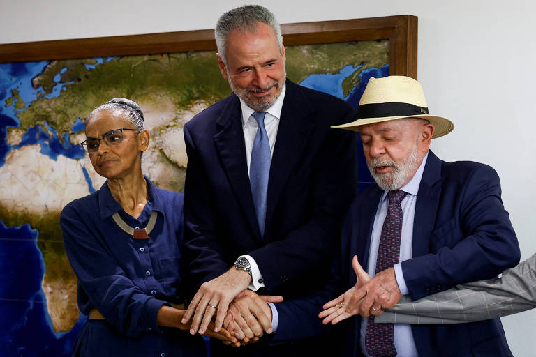 Três pessoas estão em uma reunião， com um mapa-múndi ao fundo. À esquerda， uma mulher de cabelo curto e grisalho， usando óculos e uma blusa azul escura， está com as mãos unidas. No centro， um homem de cabelo grisalho e barba， vestido com um terno escuro e gravata， sorri enquanto coloca suas mãos sobre as delas. À direita， um homem de cabelo grisalho e barba， usando um terno escuro e um chapéu claro， olhando para baixo， tenta juntar as mãos dos outros dois a de uma outra pessoa， cujo braço aparece à direita dele