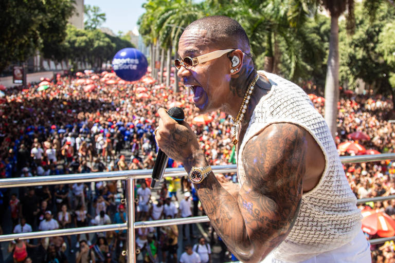 Um artista se apresenta em um palco elevado， cantando para uma grande multidão. O público é visível， com muitas pessoas reunidas em um ambiente ao ar livre， cercado por árvores. O artista está usando uma camiseta sem mangas e tem tatuagens visíveis nos braços. Há balões e bandeiras na multidão， criando um clima festivo.