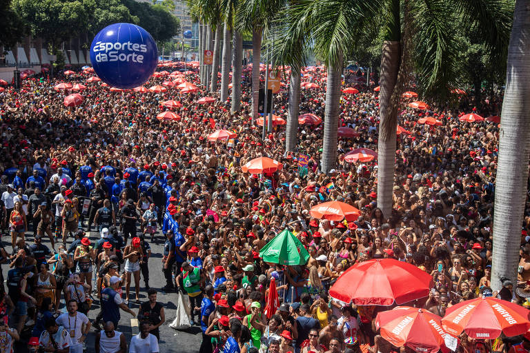 A imagem mostra uma grande multidão reunida em um evento ao ar livre， com muitas pessoas usando roupas coloridas. Há várias sombrinhas vermelhas e verdes espalhadas pela cena， além de palmeiras ao fundo. Um balão azul flutua acima da multidão， e a atmosfera parece festiva.