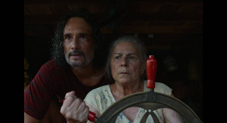 A imagem mostra dois personagens em um ambiente interno. Um homem com cabelo ondulado e barba está à esquerda, olhando para frente com uma expressão intensa. À direita, uma mulher mais velha, com cabelo grisalho e expressão séria, segura um volante de barco. O fundo é escuro, sugerindo um ambiente de barco ou casa de madeira.