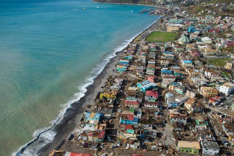 Imagem aérea de uma área costeira， mostrando uma linha de praia com ondas suaves e uma cidade ao fundo. As casas são de diferentes cores， com algumas danificadas， e há uma área verde visível. O mar é de um tom azul claro， e a costa é cercada por montanhas ao fundo.