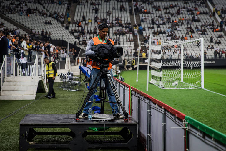 Um cinegrafista está posicionado próximo ao campo de futebol， operando uma câmera profissional montada em um tripé sobre uma plataforma. Ele veste um colete laranja de identificação e fones de ouvido. Ao fundo， vê-se parte das arquibancadas de um estádio com vários torcedores， além de um gol com rede branca. O cinegrafista está focado na captação de imagens durante um evento esportivo.







