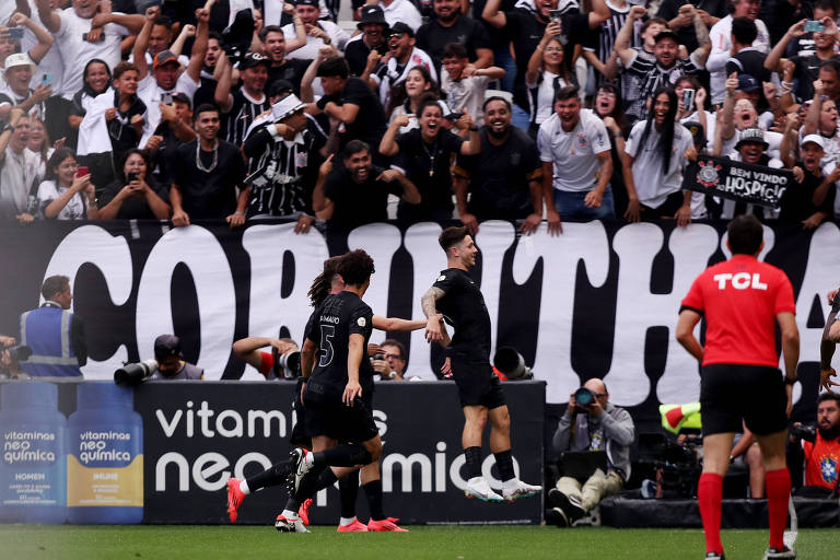 A imagem mostra um momento de celebração de um gol do time de futebol Corinthians. Jogadores estão se abraçando em campo， enquanto a torcida ao fundo está animada， com muitos torcedores vestindo camisas listradas em preto e branco. Um grande banner com a palavra 039;CORINTHIANS039; é visível atrás da torcida. Um árbitro em uniforme vermelho está próximo aos jogadores.