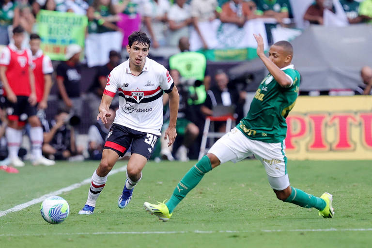 A imagem mostra um momento de um jogo de futebol， onde um jogador com a camisa do São Paulo， número 39， está correndo em direção à bola， que está no chão. Ele é perseguido por um jogador do time adversário， que está vestindo uma camisa verde. Ao fundo， há torcedores e banners visíveis， indicando um ambiente de estádio.