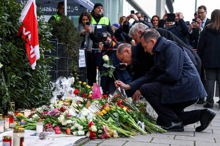 A imagem mostra um grupo de pessoas em um ato de homenagem， com três homens ajoelhados em frente a um monte de flores e velas. Eles estão colocando flores em um memorial， que é cercado por uma cerca baixa. Ao fundo， há várias pessoas observando e registrando o momento com câmeras. Um dos homens está segurando uma rosa branca. O ambiente parece ser ao ar livre， com um edifício visível ao fundo.