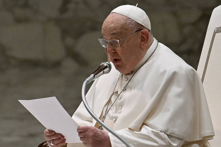 O Papa Francisco está em pé， segurando um papel e falando em um microfone. Ele usa uma vestimenta branca e um pálio， com uma mitra branca na cabeça. O fundo é neutro e desfocado， sugerindo um ambiente formal.