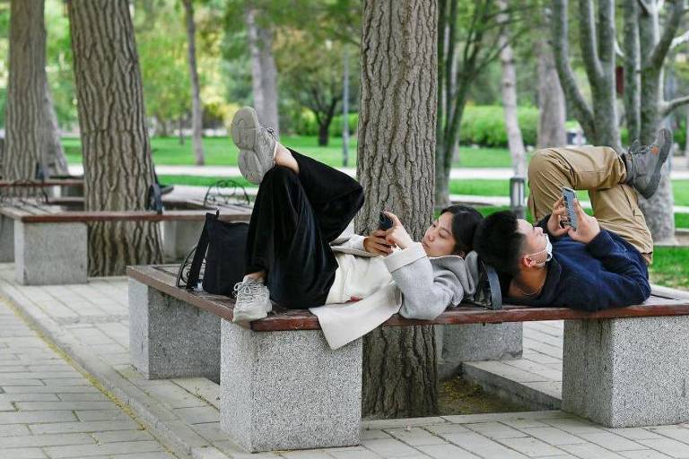 A imagem mostra duas pessoas deitadas em um banco de parque， com as pernas para cima e usando seus celulares. O ambiente é arborizado， com árvores ao fundo e um gramado verde. As pessoas estão vestidas de forma casual， uma delas usa um casaco claro e a outra um suéter escuro.