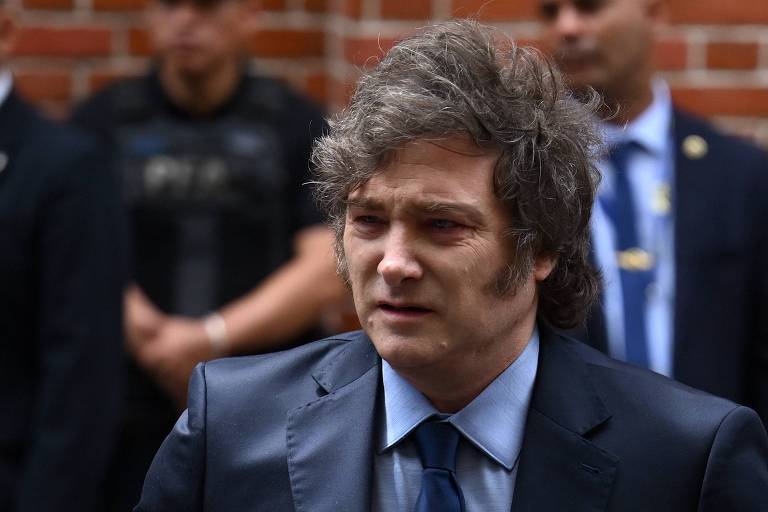 Um homem com cabelo desgrenhado e um terno escuro está em primeiro plano， parecendo preocupado. Ele usa uma gravata azul e está em um ambiente externo， com um muro de tijolos ao fundo. Ao fundo， há várias pessoas em trajes de segurança， observando a cena.