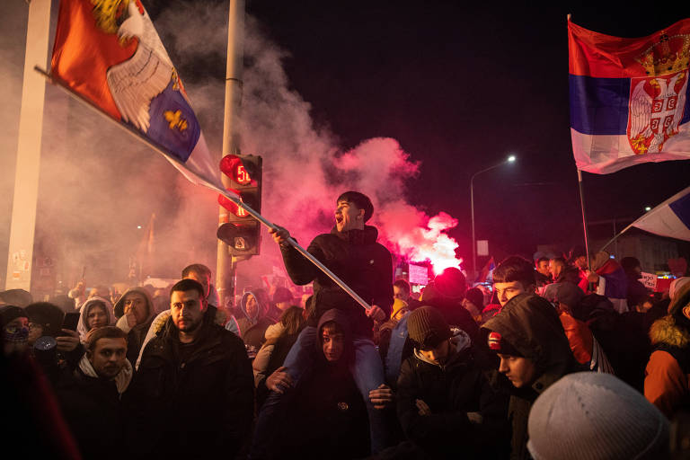 A imagem mostra uma multidão em um protesto à noite， com fumaça e sinalizadores. Um homem está em cima de outra pessoa， segurando uma bandeira. Várias pessoas ao redor estão com expressões de entusiasmo ou determinação， e algumas também seguram bandeiras. O ambiente é iluminado por luzes e há um sinal de trânsito visível ao fundo.