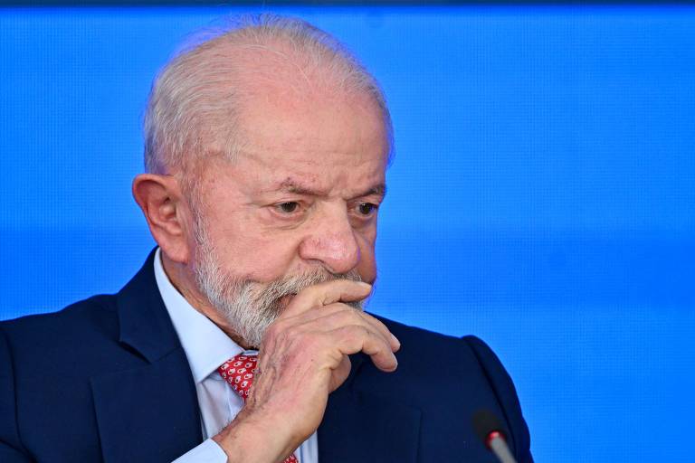 Um homem com cabelo grisalho e barba， vestindo um terno escuro e uma gravata vermelha com padrões， está sentado em uma mesa. Ele parece pensativo， com a mão cobrindo a boca e o olhar voltado para baixo. O fundo é azul， sugerindo um ambiente de conferência ou evento formal.