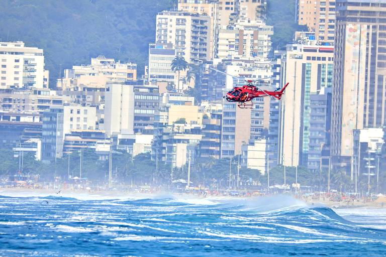 A imagem mostra um helicóptero vermelho voando sobre o mar, com ondas visíveis na parte inferior. Ao fundo, há uma linha de prédios altos e modernos, e uma praia com pessoas visíveis na areia. O céu está claro, e a cena transmite uma sensação de atividade e movimento.