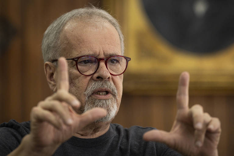 Um homem de cabelos grisalhos e barba， usando óculos de armação vermelha， está gesticulando com as mãos enquanto fala. Ele veste uma camiseta escura e parece estar em um ambiente interno， com uma parede de madeira ao fundo e um quadro emoldurado visível.