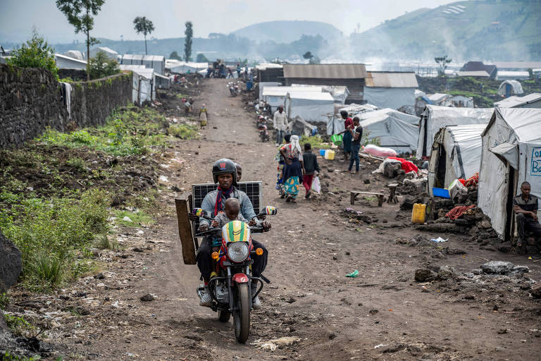 A imagem mostra um acampamento de refugiados com várias barracas brancas ao longo de um caminho de terra. Um homem em uma motocicleta transporta uma criança e um grande painel. Ao fundo， há pessoas caminhando e interagindo， com algumas barracas e árvores visíveis. O ambiente parece ser rural， com montanhas ao longe.