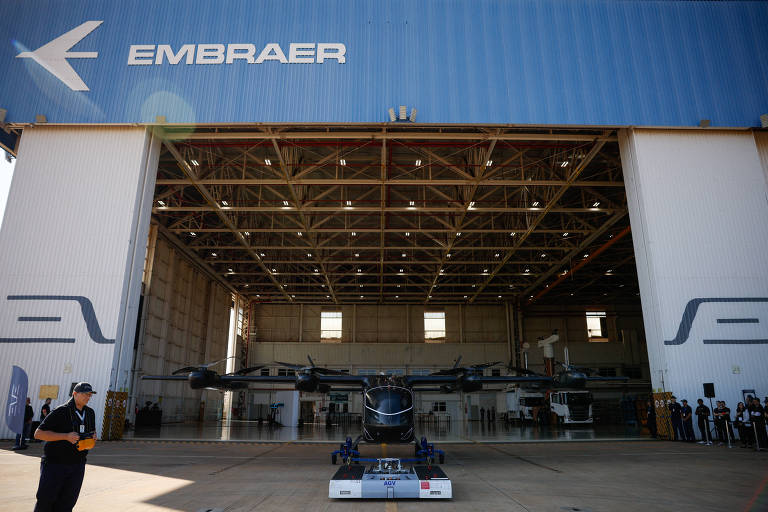 A imagem mostra o hangar da Embraer， com a entrada ampla e um veículo aéreo não tripulado em destaque na frente. O hangar é grande， com uma estrutura metálica visível e o logotipo da Embraer na parte superior. Há pessoas em pé ao lado do veículo， observando.