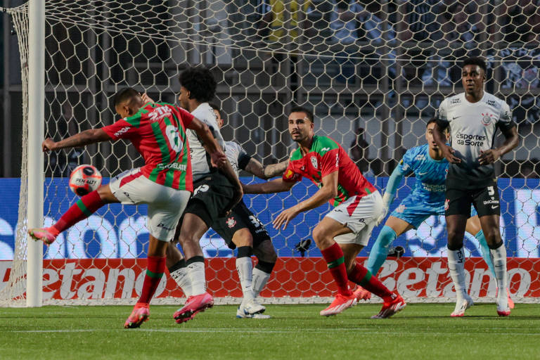 A imagem mostra um momento de um jogo de futebol, onde um jogador em destaque, vestindo uma camisa vermelha e verde, está prestes a chutar a bola. Outros jogadores estão posicionados ao redor, incluindo um goleiro em uniforme azul e outros jogadores em camisas brancas e pretas. A rede do gol está visível ao fundo, e há uma grande quantidade de torcedores nas arquibancadas.