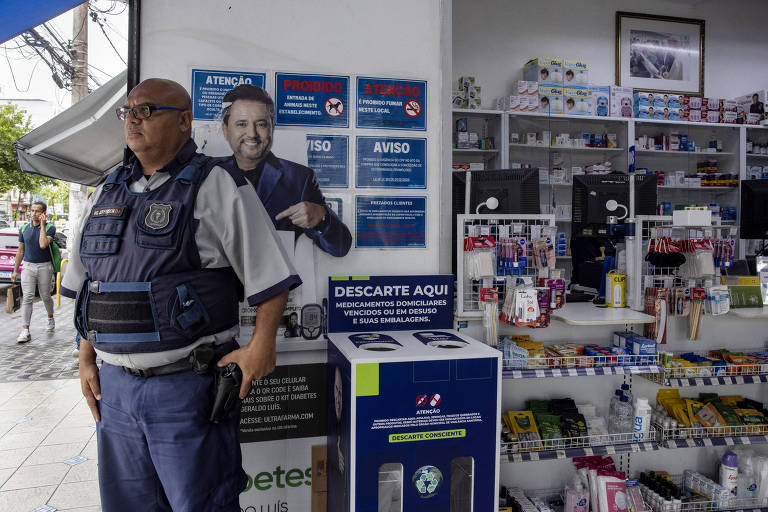 A imagem mostra um policial de pé em frente a uma farmácia. Ao fundo， há uma prateleira com diversos produtos farmacêuticos. Na parede， estão afixados vários avisos e um cartaz com a imagem de um homem sorridente. Há também uma caixa de descarte de medicamentos na parte inferior direita da imagem.