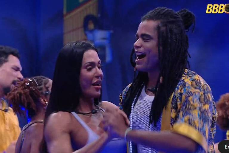 A imagem mostra um grupo de pessoas dançando em um ambiente festivo. No primeiro plano, um homem com cabelo longo e dreadlocks está sorrindo e segurando a mão de uma mulher com cabelo longo e liso.  Ao fundo, outras pessoas também estão dançando, com um homem usando uma camisa amarela e uma mulher com cabelo cacheado. O cenário é iluminado com luzes azuis e há elementos decorativos que sugerem um ambiente de festa.