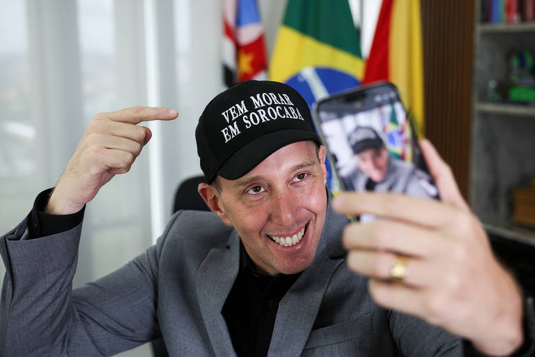 Um homem está tirando uma selfie enquanto usa um boné preto com a frase 039;VEM MORAR EM SOROCABA039;. Ele está sorrindo e apontando para o boné. Ao fundo， há bandeiras do Brasil e do estado de São Paulo， além de uma estante com livros.