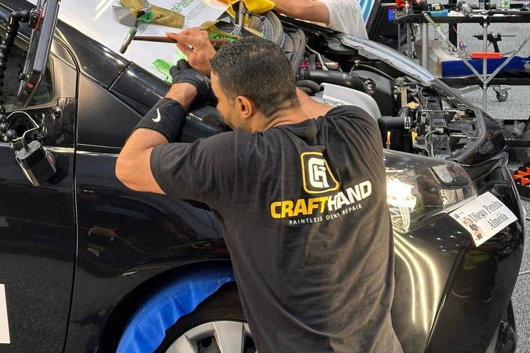 A imagem mostra um evento de competição onde várias pessoas estão trabalhando em veículos. Cada participante está aplicando um filme em um carro preto， utilizando ferramentas como espátulas e panos. O ambiente é bem iluminado， com várias luzes direcionadas para os carros. Ao fundo， há uma tela grande e outras pessoas assistindo ao evento.